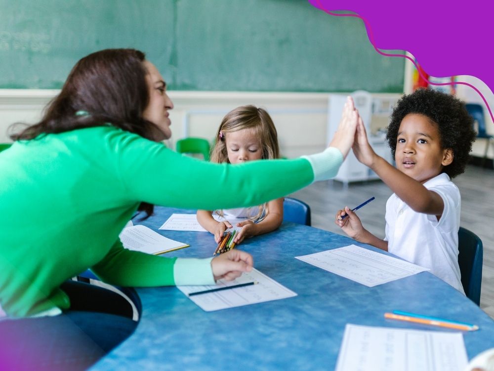 Professora comemorando com um aluno com em um gesto que chamamos “bate aqui”, enquanto outra criança desenha, representando um ambiente positivo e de apoio aos alunos com dificuldades de aprendizagem nos anos iniciais.
