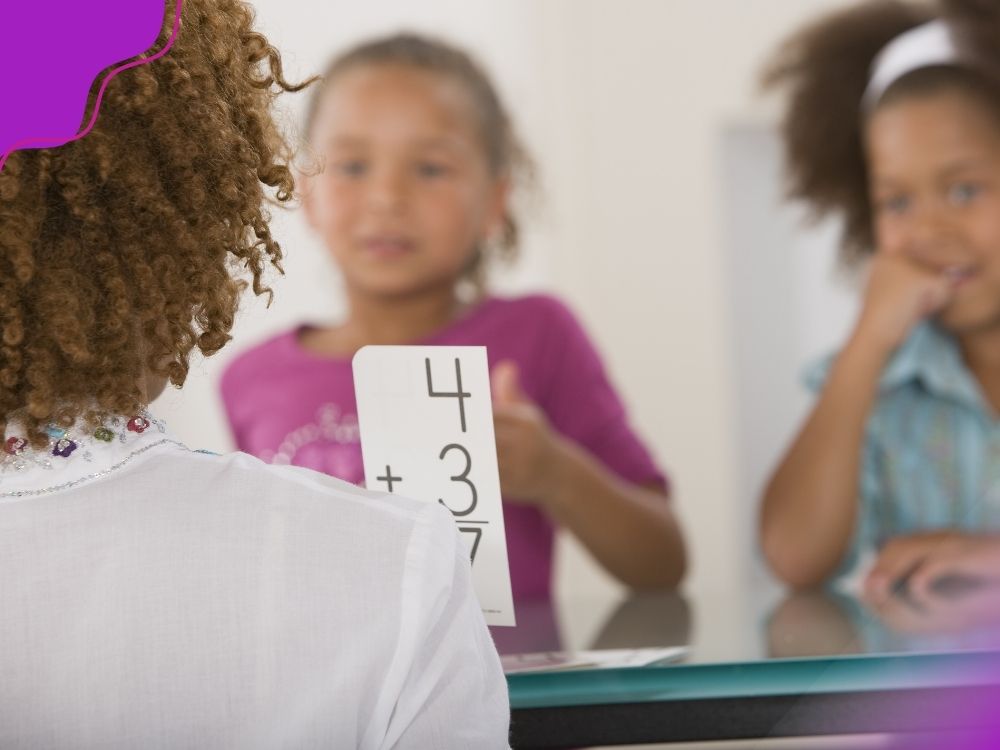 Professora segurando um flashcard com uma operação matemática básica (4+3) para ilustrar uma estratégia para intervenção pedagógica de matemática.
