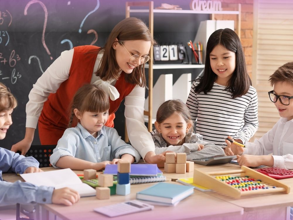 Alunos com expressão de felicidade e motivação, vários materiais concretos na mesa escolar e professora realizando intervenções. A imagem ilustra como os professores podem ajudar os alunos a terem uma relação positiva com a matemática, superando a ansiedade.