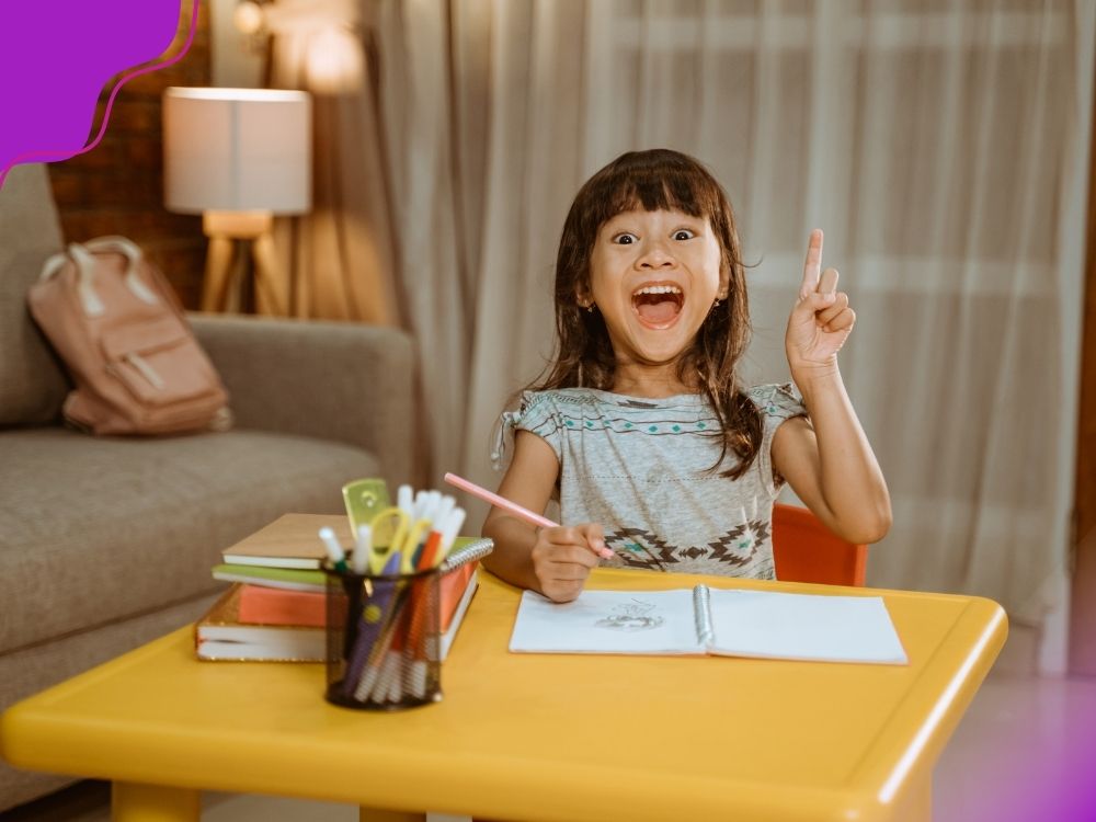 Menina sorridente sentada a uma mesa amarela com caderno aberto, segurando um lápis e levantando o dedo indicador como se tivesse tido uma ideia. Ao lado, há livros empilhados e um porta-canetas. A imagem ilustra a importância da metacognição para desenvolver autonomia dos alunos.