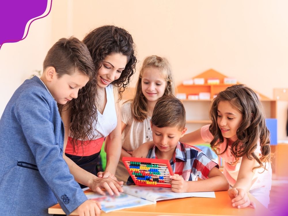 Professora interagindo com um grupo de quatro crianças em uma sala de aula. Uma delas segura um ábaco colorido, enquanto os demais observam com atenção e curiosidade. Há um livro didático sobre a mesa. A imagem ilustra a importância das intervenções para desenvolver o senso numérico.